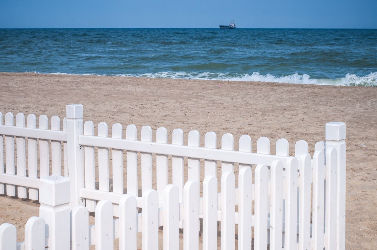 Elegant and simple white wooden fence on beach sand with sea surf background. Minimal summer seascape with blurred vessel on horizon. Summer sunny day. Coastal landscape with white striped fence